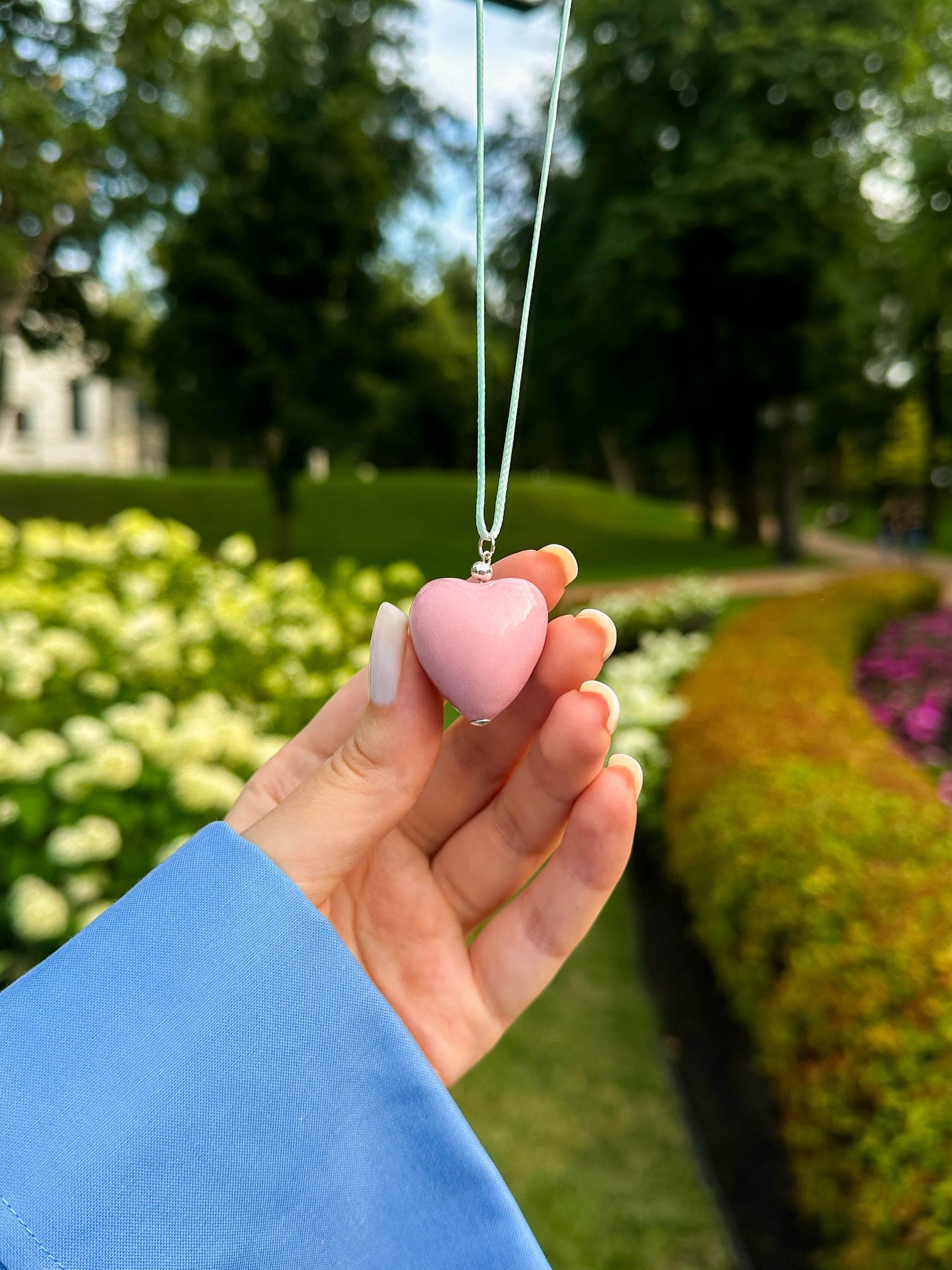 A pink heart-shaped pendant made with ashes, held between two fingers in a garden